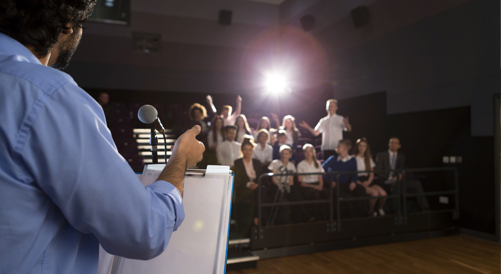 man at a lectern address a man in the audience who is standing up with arms raised