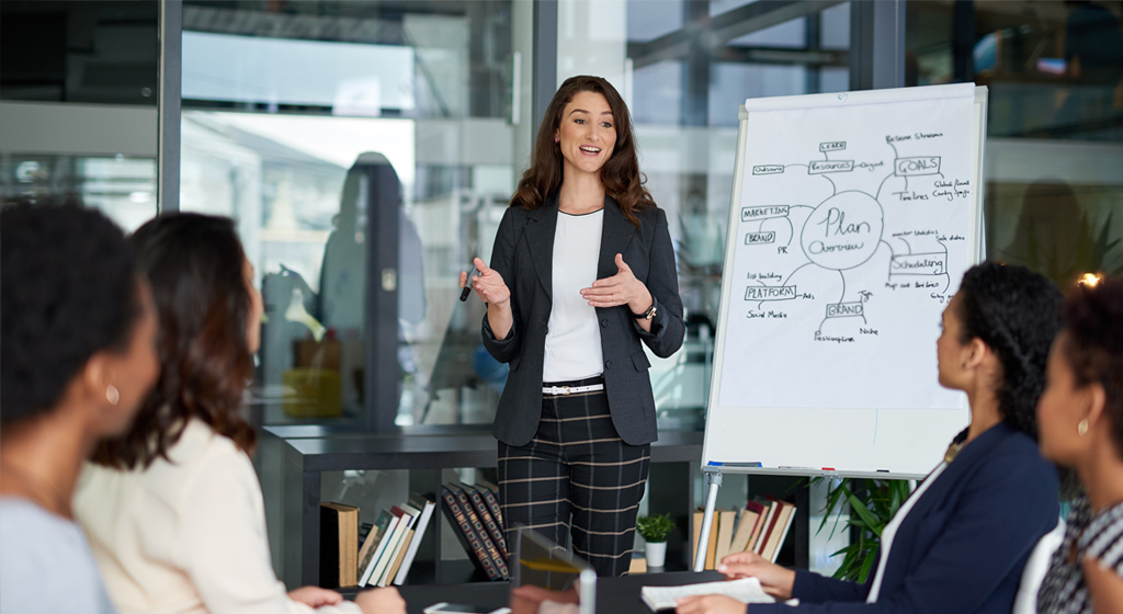 woman standing in front of a white board talking to a group of people seated at a conference table.