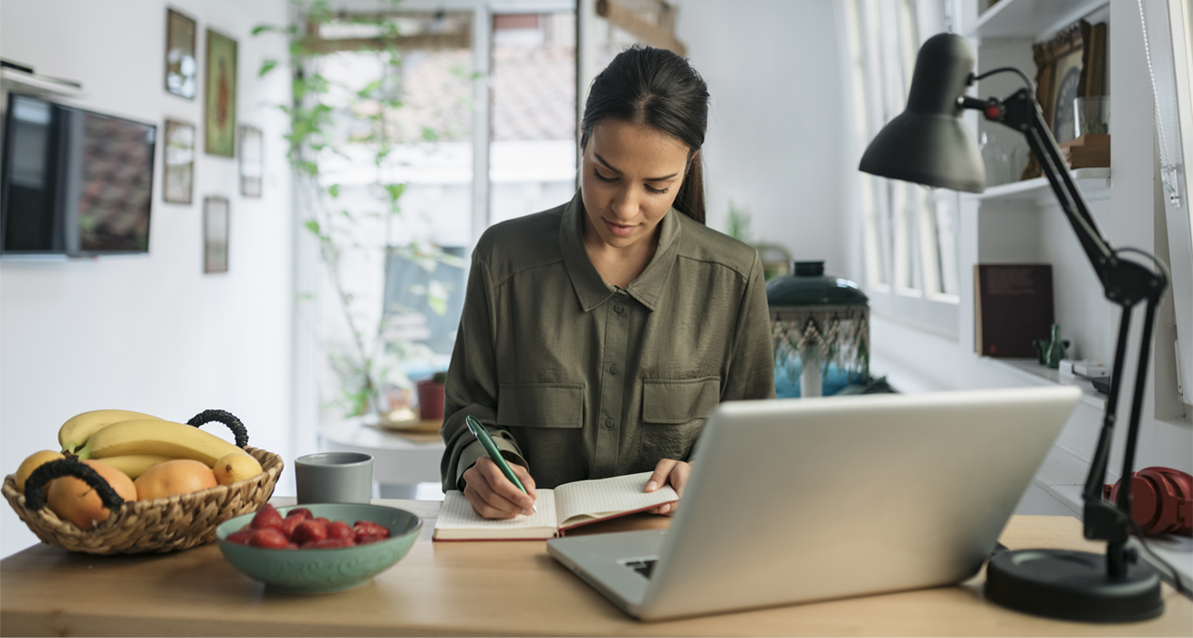 woman at a table writing in a journal in front of a laptop computer.