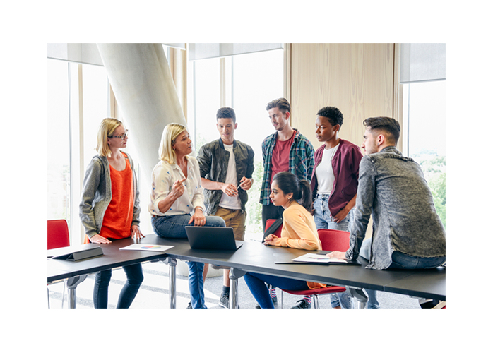 several college students standing an sitting at two tables looking and listening to an instructor who is sitting on one of the tables