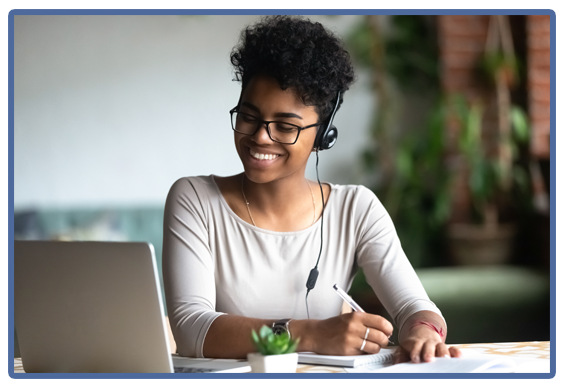 woman writing in a book wearing a headset, smiling while looking at laptop computer screen