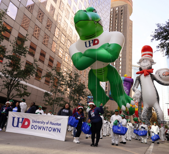 Ed-U-Gator parade balloon, adults holding a University of Houston-Downtown banner, and a Cat in the Hat parade balloon at the heb thanksgiving day parade