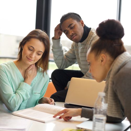 Three people sit at a table with a textbook open between them, studying the pages together.