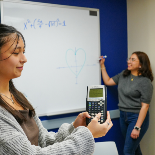 A student holds a calculator with a heart on it while a second student works a math equation on a whiteboard in the W.I. Dykes Library at the University of Houston-Downtown