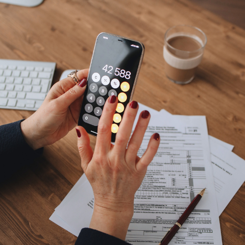 An adult holds a phone while typing numbers on the calculator app. A desk has tax papers, keyboard, and cup of coffee on it. 