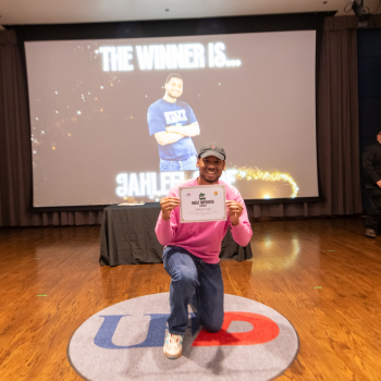 A UHD student athlete kneels while holding a UHD sports and fitness awards certificate